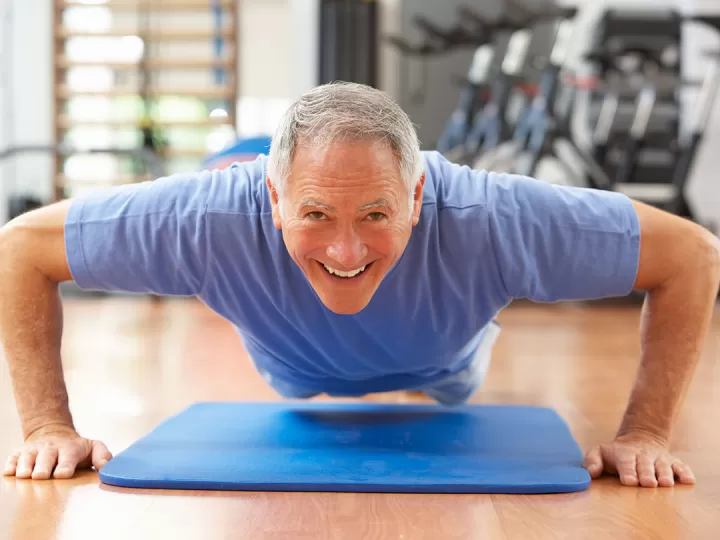 An older gentleman with grey hair and a blue t shirt is hovering in a pushup over a blue mat in a gym, and smiling at the camera. 
